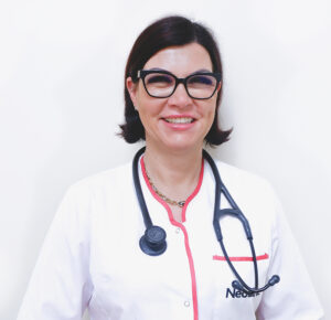 Lekarka Katarzyna Zabłocka stoi, ma na sobie biały fartuch, na szyi stetoskop, uśmiecha się