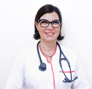 Lekarka Katarzyna Zabłocka stoi, ma na sobie biały fartuch, na szyi stetoskop, uśmiecha się