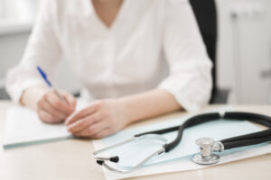Lekarka wypełniająca druki , siedzi przy biurku, trzyma długopis w prawej ręce, na pierwszy planie leży stetoskop